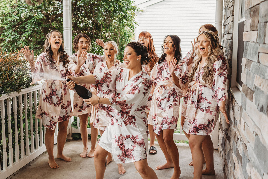 bride popping champagne outdoors with bridesmaids wearing matching robes