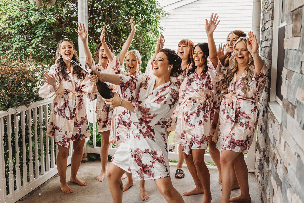 bride popping champagne outdoors with bridesmaids wearing matching robes